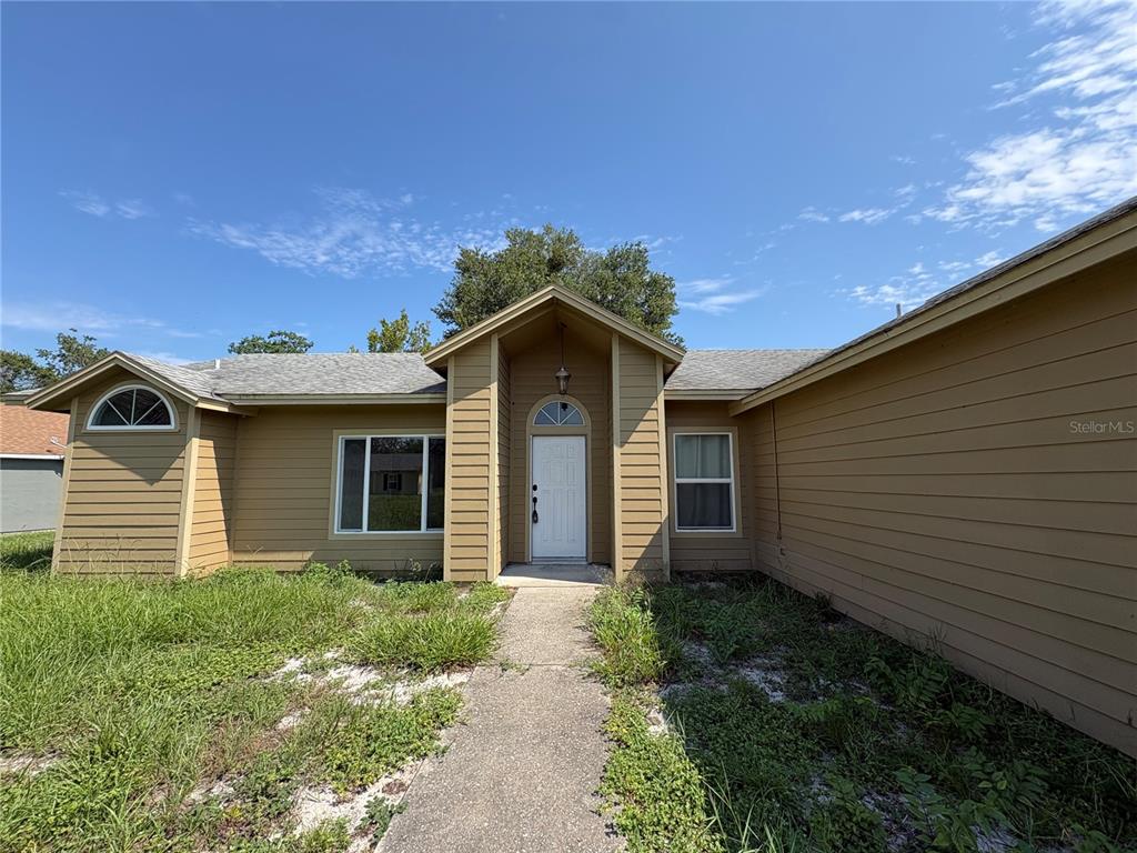 A tan, single-story house with horizontal siding, a gabled entryway, and an overgrown front yard — 2302 BELEN DRIVE, DELTONA, FL