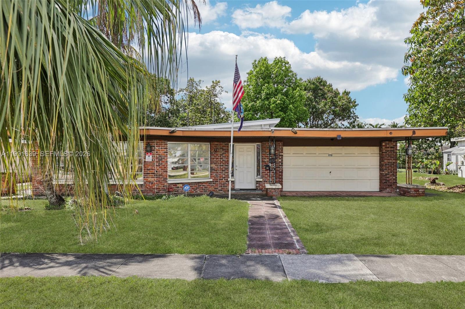 A single-story brick house features a white garage door, a front walkway, and an American flag — 272 W 46th St, Hialeah FL 33012, HIALEAH, FL