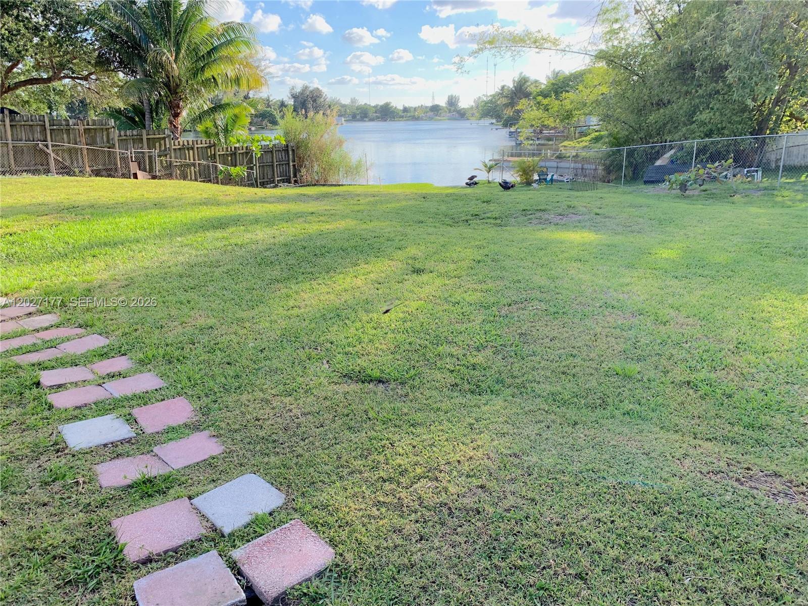 A grassy backyard features a stone pathway leading toward a lake and wooden fence — 3300 SW 32nd Ave, West Park FL 33023, WEST PARK, FL
