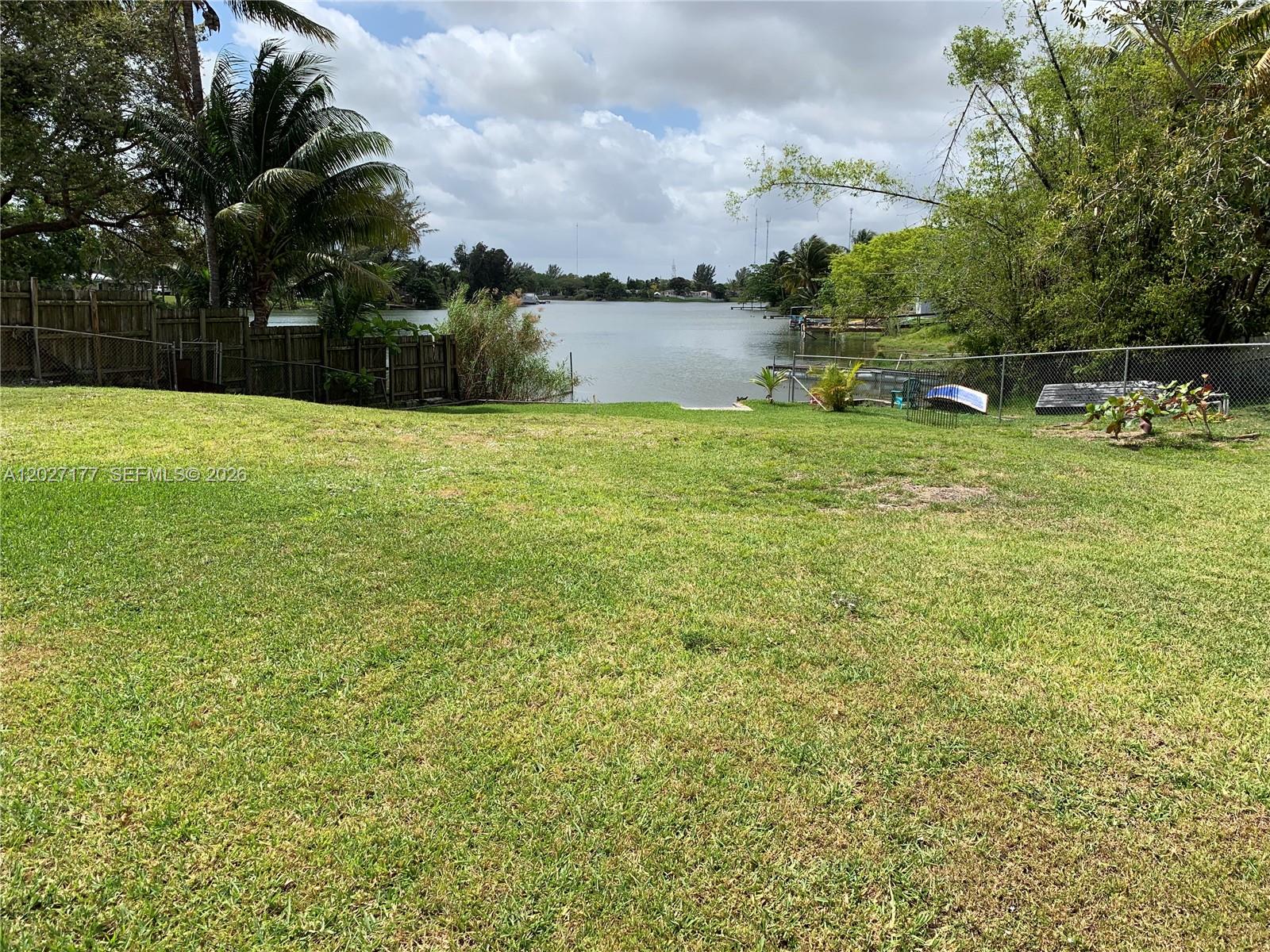 A grassy backyard leads to a waterfront view with a wooden fence and trees — 3300 SW 32nd Ave, West Park FL 33023, WEST PARK, FL