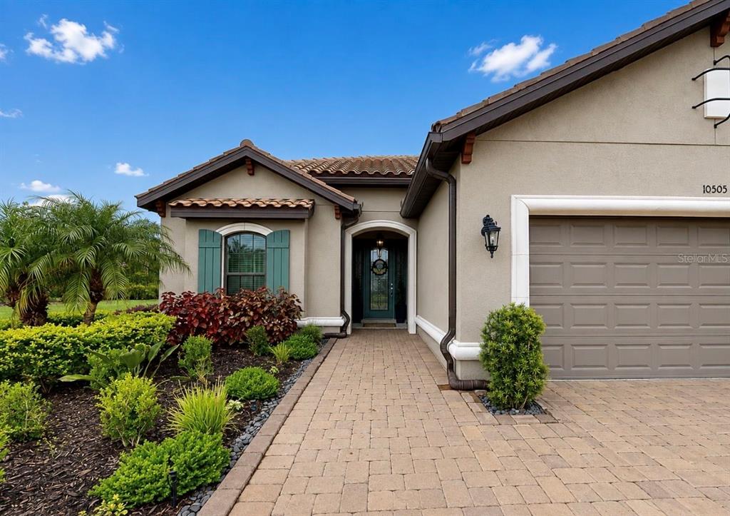 A beige house exterior features a paved walkway, a garage, and green shutters — 10505 WICKER PARK PLACE, PALMETTO, FL