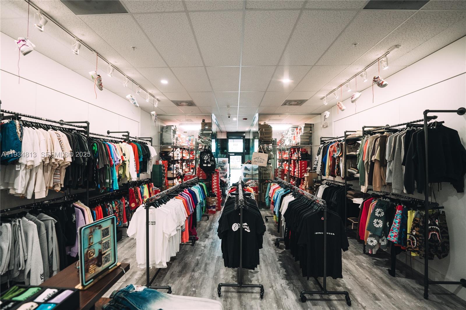 A retail clothing store features rows of hanging apparel, shelves of shoeboxes, and ceiling track lighting — 12700 SW 128th St #106, Miami FL 33186, MIAMI, FL