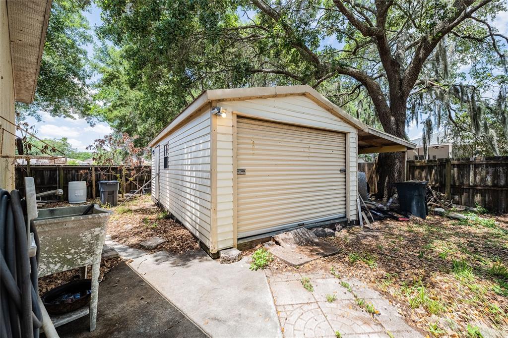 A detached shed with a roll-up garage door sits in a backyard with trees — 427 LARK COURT, KISSIMMEE, FL