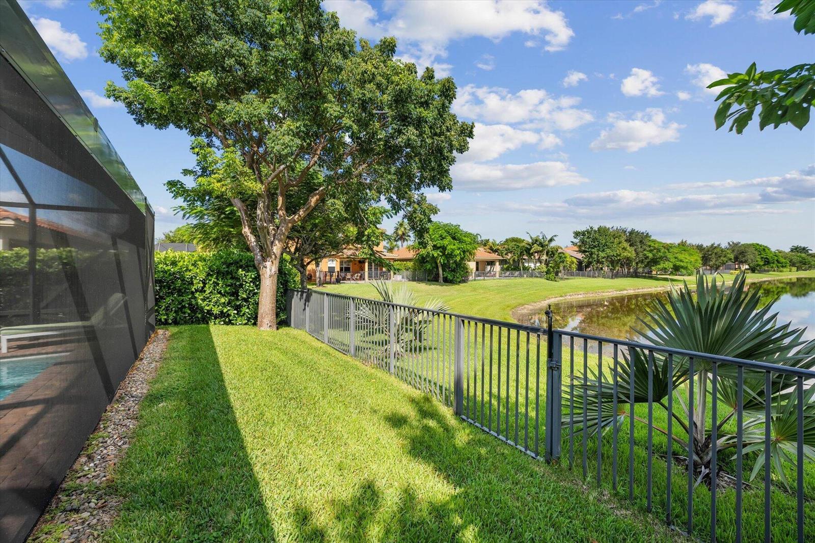 A fenced backyard features a green lawn, a large tree, and a view of a pond — 10731 Ivanhoe Lane, Wellington FL 33414, WELLINGTON, FL