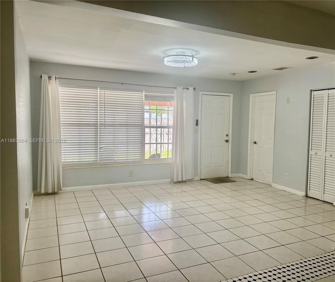 A living room features white tile flooring, a large window with blinds, and white doors — 2016 NE 26th St # 2, Wilton Manors FL 33305, WILTON MANORS, FL