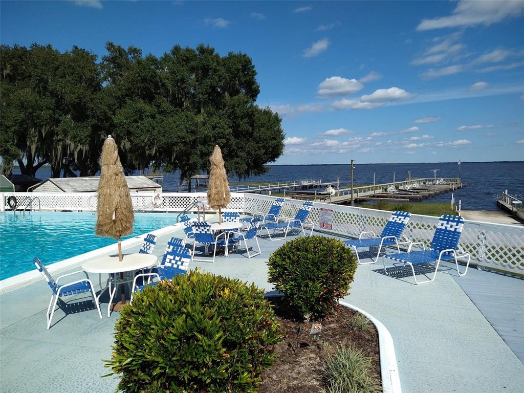 A swimming pool deck with lounge chairs, tables, and umbrellas overlooks a lake and dock — 8454 BREEZE HILL DRIVE, LAKE WALES, FL