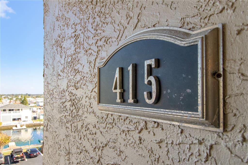 A close-up view of a house number plaque reading 415 mounted on a textured exterior wall — 6035 SEA RANCH DRIVE UNIT 415, HUDSON, FL
