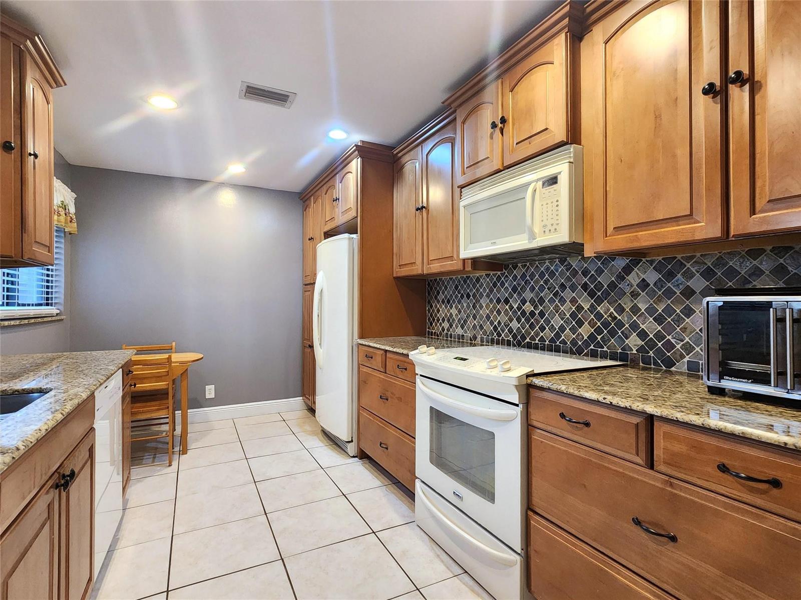 A kitchen features wood cabinets, granite countertops, a tiled backsplash, and white appliances — 1572 SW 23rd Terrace, Deerfield Beach FL 33442, DEERFIELD BEACH, FL