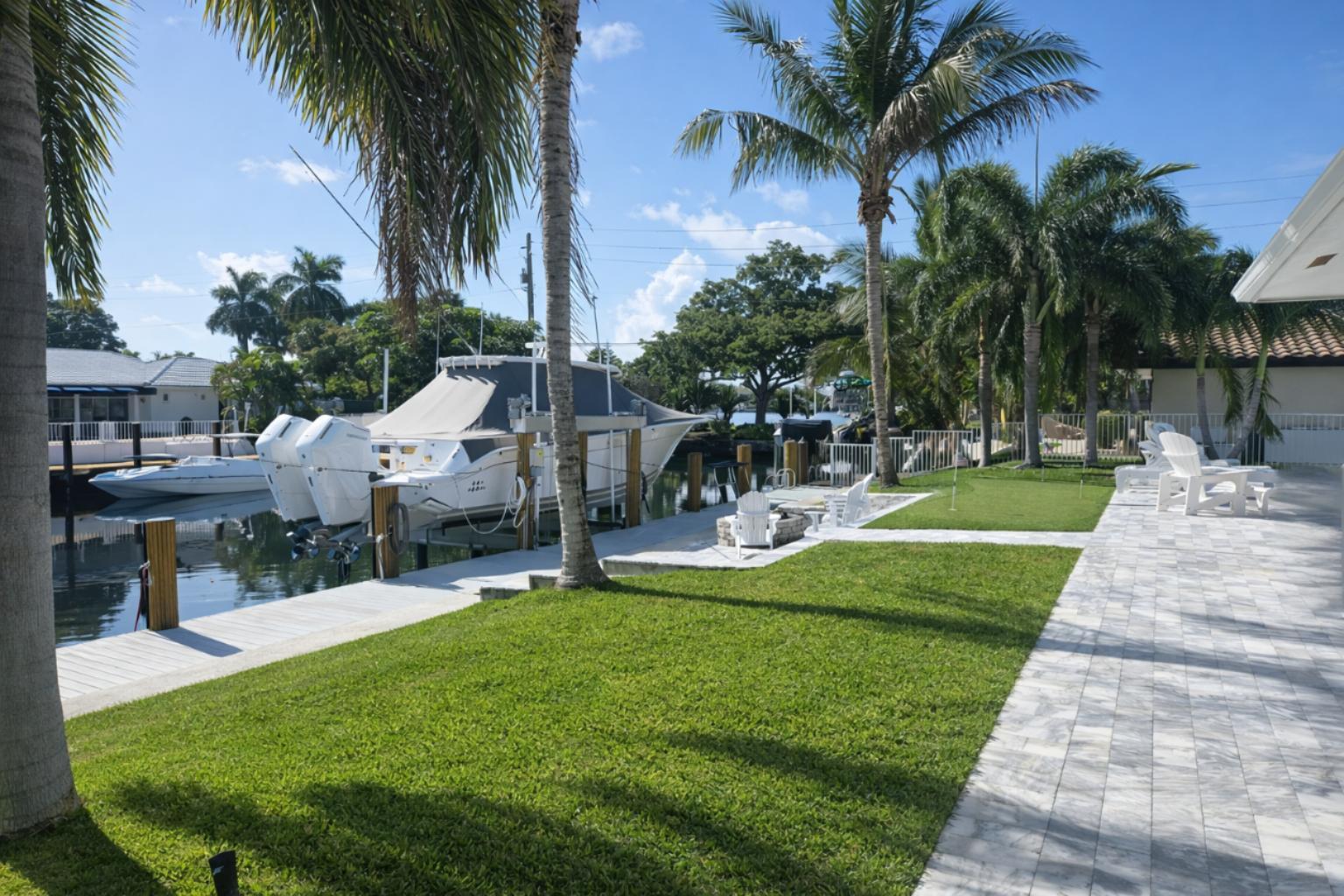 A backyard with a lawn, stone patio, palm trees, and a boat docked on a canal — 3910 NE 26th Avenue, Lighthouse Point FL 33064, LIGHTHOUSE POINT, FL