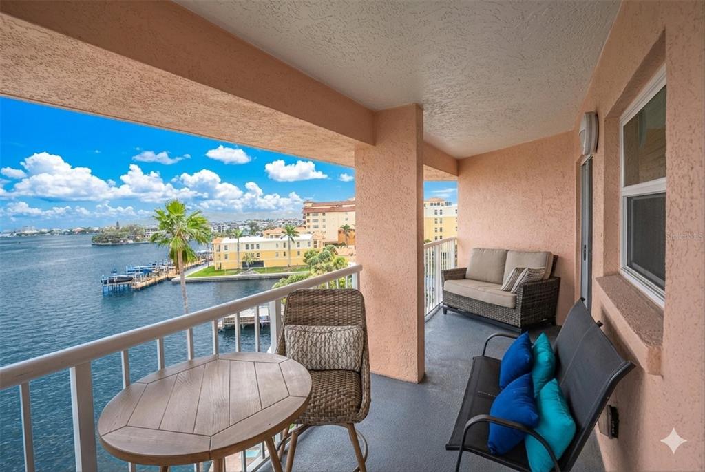 A covered balcony with outdoor furniture overlooks a bay with a dock and distant buildings — 5445 GULF BOULEVARD UNIT PH1, ST PETE BEACH, FL
