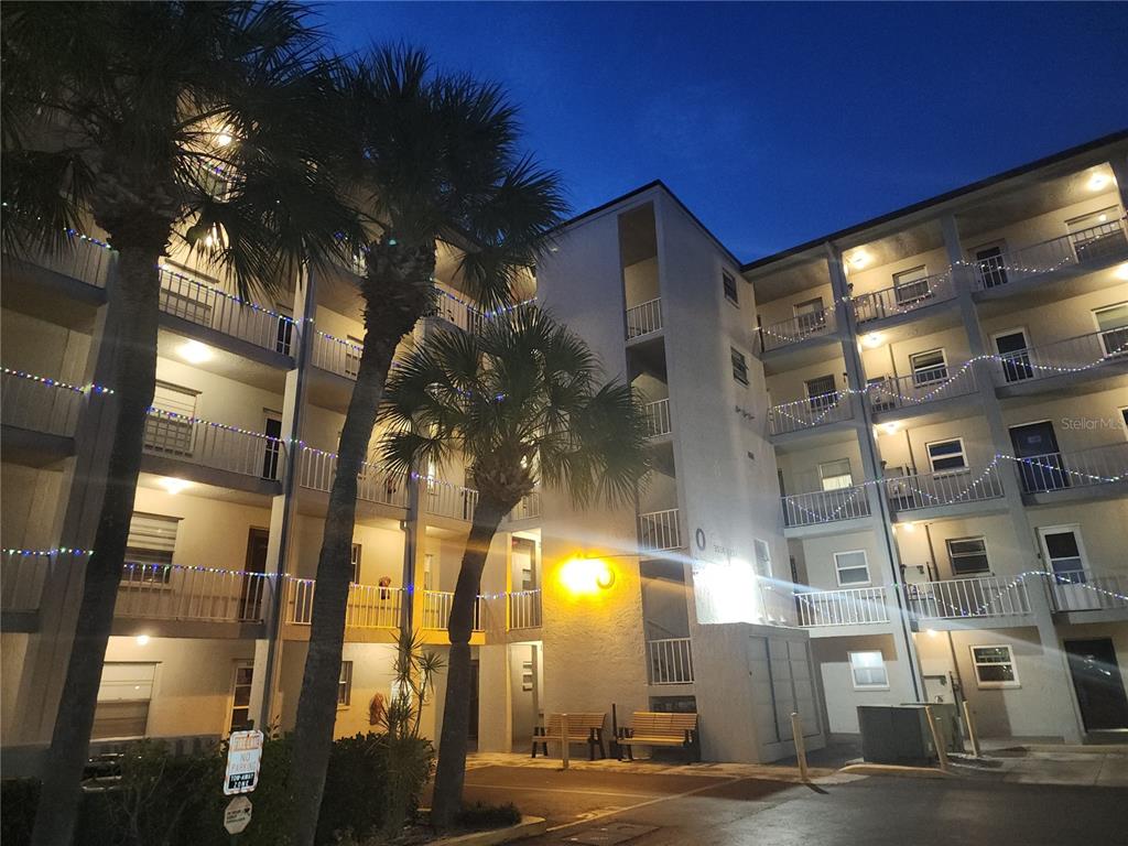 An exterior view of a multi-story apartment building at dusk with palm trees and lights — 3064 LAKE BAYSHORE DRIVE UNIT 115, BRADENTON, FL