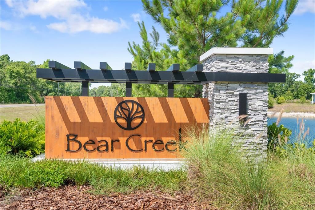 A stone and metal monument sign for Bear Creek stands outdoors near a pond — 12003 GRIZZLY LANE, NEW PORT RICHEY, FL