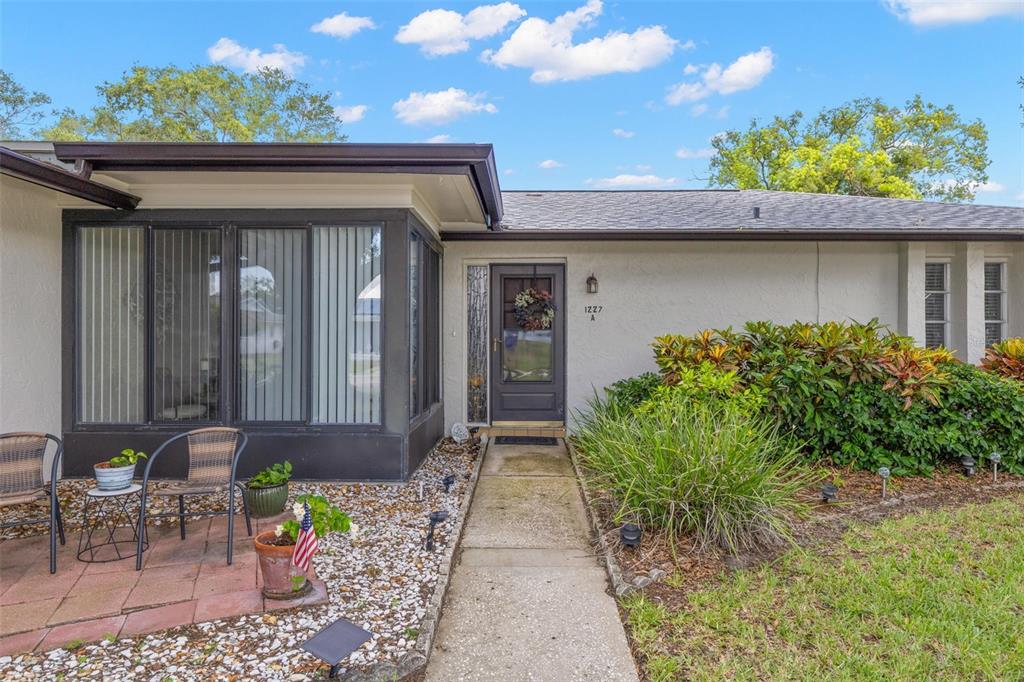 The exterior of a single-story house features a concrete walkway, a glass door, and landscaping — 1227 QUEEN ANNE DRIVE UNIT A, PALM HARBOR, FL