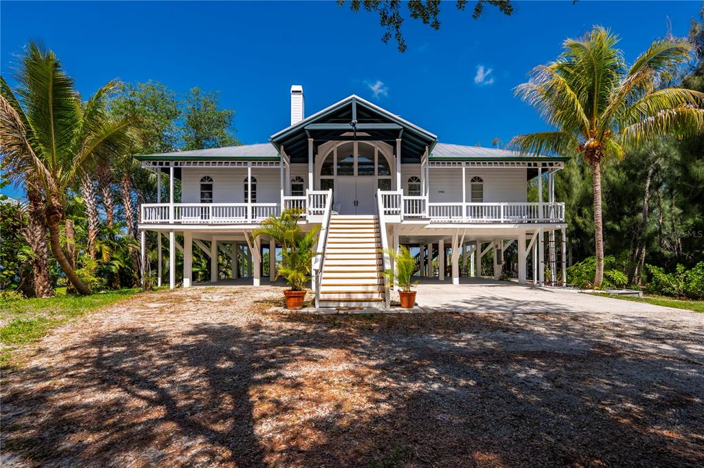 A white stilt house with a wraparound porch and central staircase stands on a sandy lot — 27414 MISTY AVENUE, PUNTA GORDA, FL