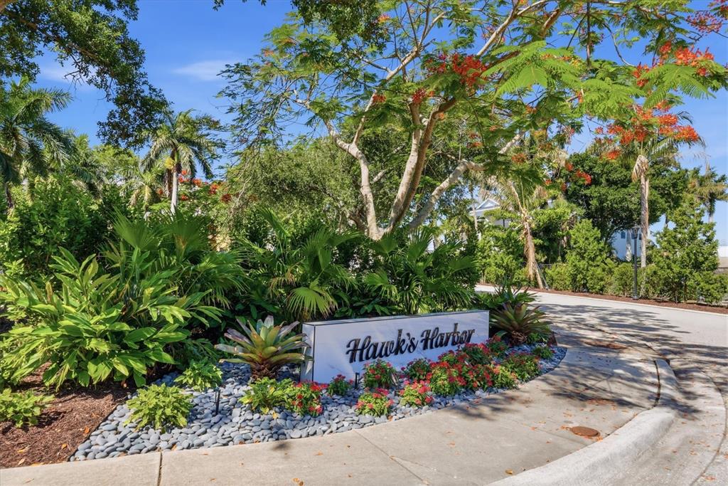 A stone sign reading Hawk's Harbor sits in a landscaped garden beside a paved road — 7107 HAWKS HARBOR CIRCLE, BRADENTON, FL