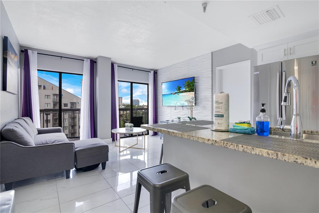 A modern living room and kitchen area with tile floors, a balcony, and a television — 6165 CARRIER DRIVE UNIT 2605, ORLANDO, FL