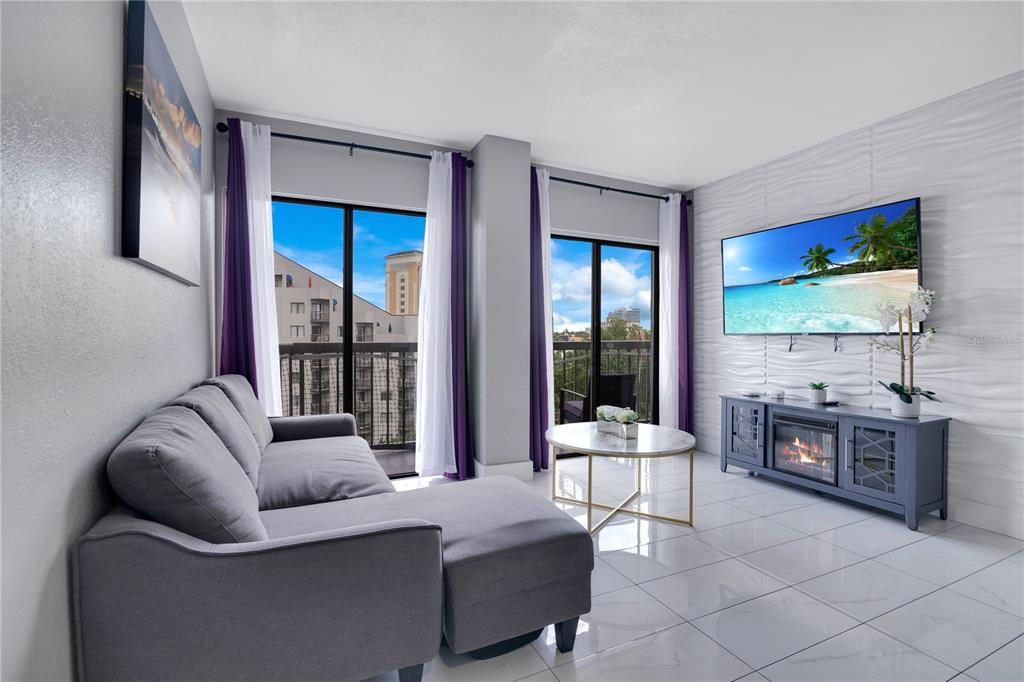 A living room features a gray sectional sofa, tiled floors, and a wall-mounted television — 6165 CARRIER DRIVE UNIT 2605, ORLANDO, FL