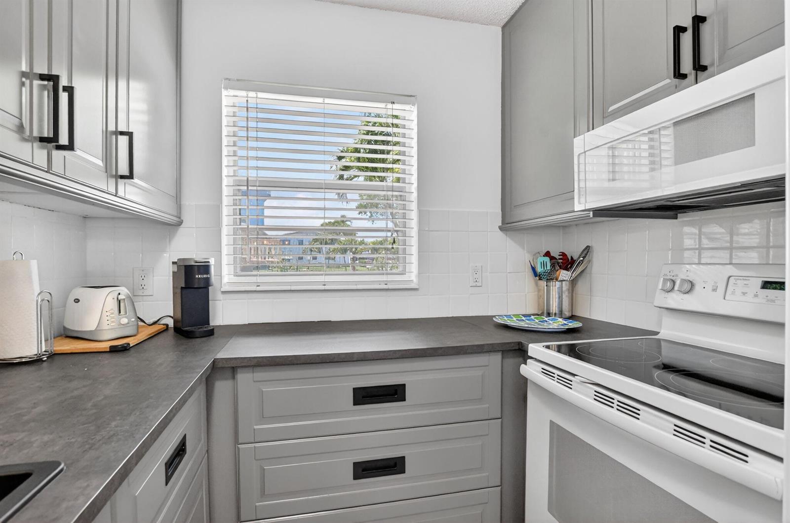 A kitchen features gray cabinets, dark countertops, white tile backsplash, and a window with blinds — 492 W Tilford, Deerfield Beach FL 33442, DEERFIELD BEACH, FL