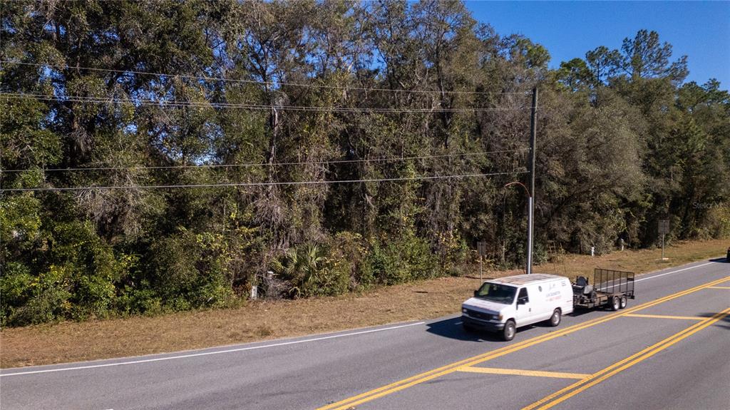 A white work van with a trailer drives along a road beside a wooded lot — 815 NE 145TH AVENUE ROAD, SILVER SPRINGS, FL