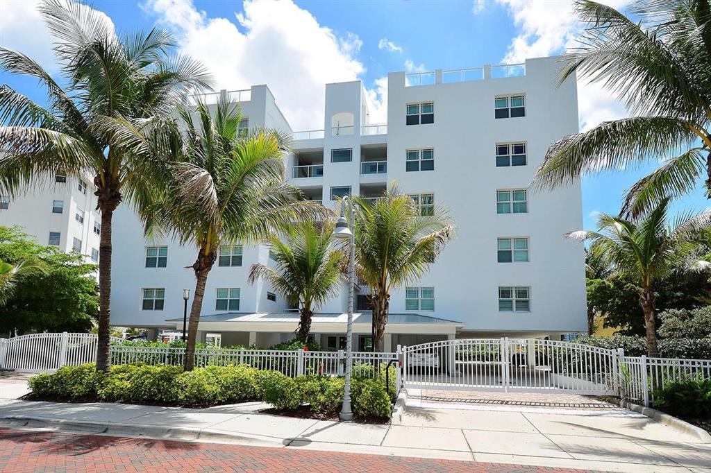A white multi-story apartment building stands behind a white metal fence and palm trees — 650 GOLDEN GATE POINT UNIT 602, SARASOTA, FL