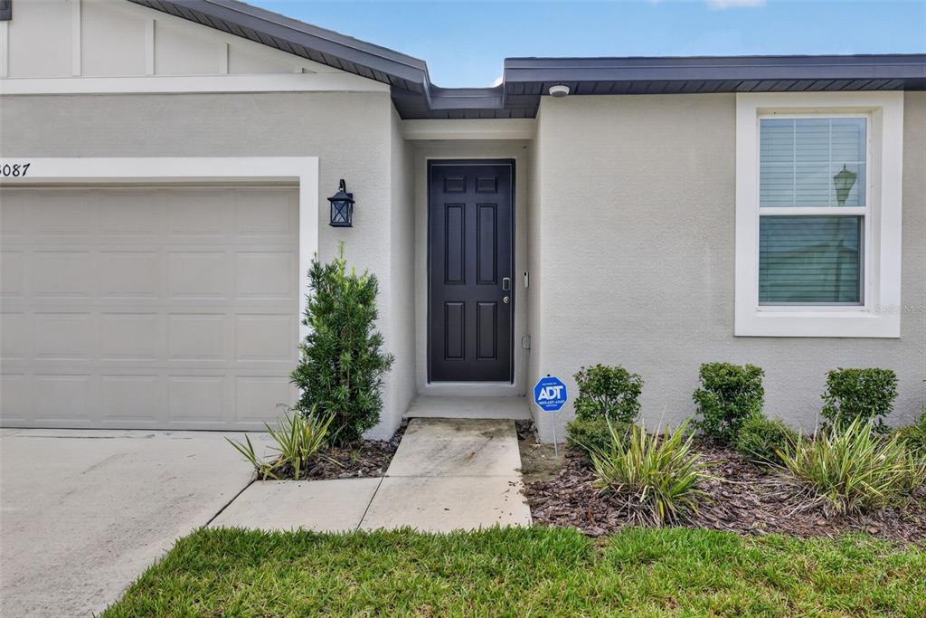 A gray house exterior features a dark front door, a garage, and a small walkway — 3087 FORREST PLUM COURT, ZEPHYRHILLS, FL