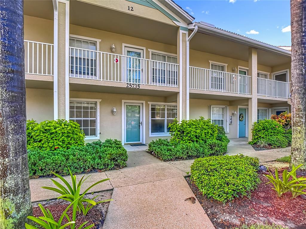 A two-story apartment building exterior features tan stucco walls, white railings, and manicured green landscaping — 25735 LAKE AMELIA WAY UNIT 103, BONITA SPRINGS, FL