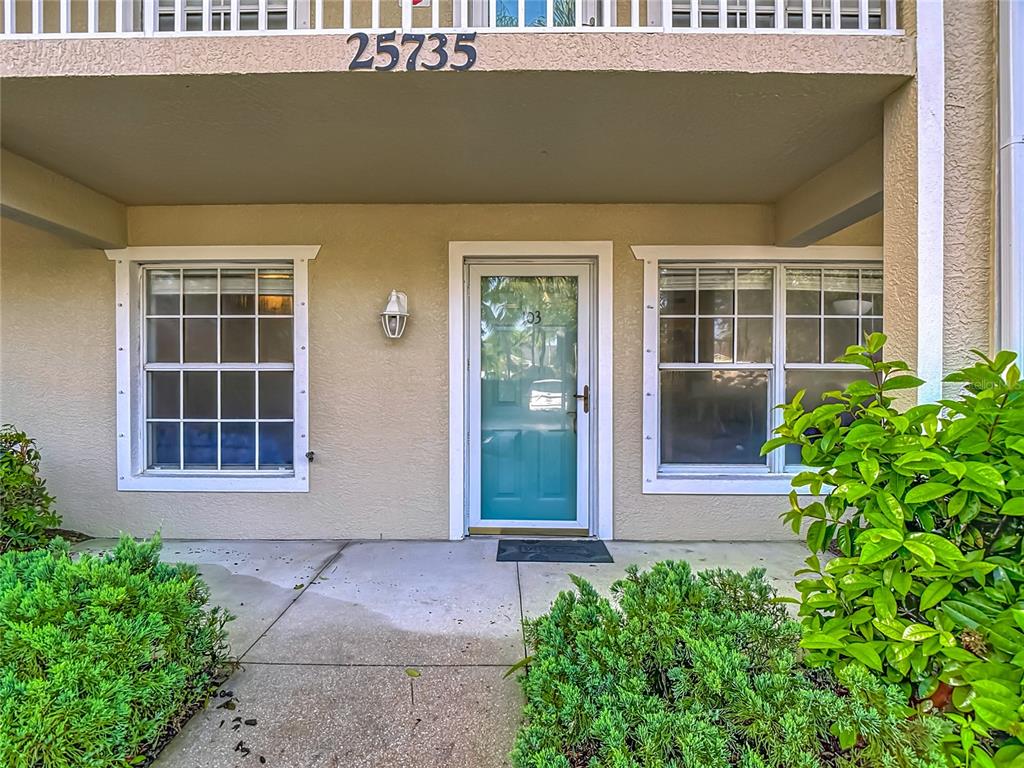A ground-level building entrance features a teal front door, two windows, and surrounding landscaping — 25735 LAKE AMELIA WAY UNIT 103, BONITA SPRINGS, FL