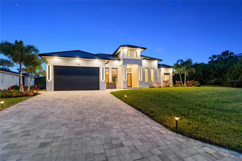 A modern single-story house with a paved driveway, dark garage door, and illuminated exterior — 13125 IRWIN DRIVE, PORT CHARLOTTE, FL