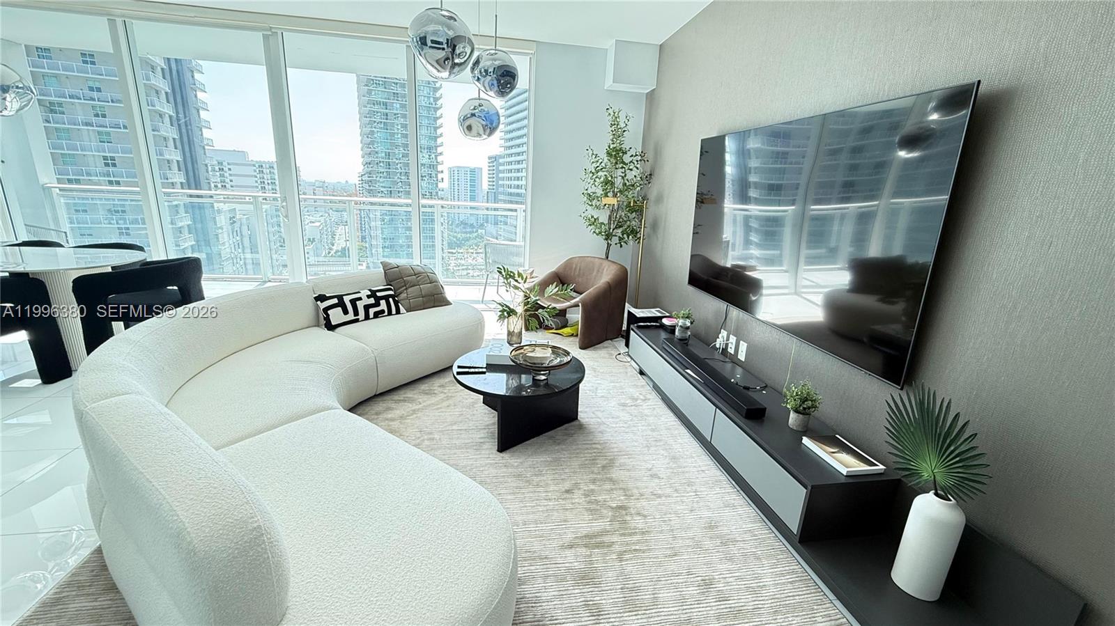 A modern living room features a curved white sofa, a wall-mounted television, and floor-to-ceiling windows — 1080 Brickell Ave # 1801, Miami FL 33131, MIAMI, FL