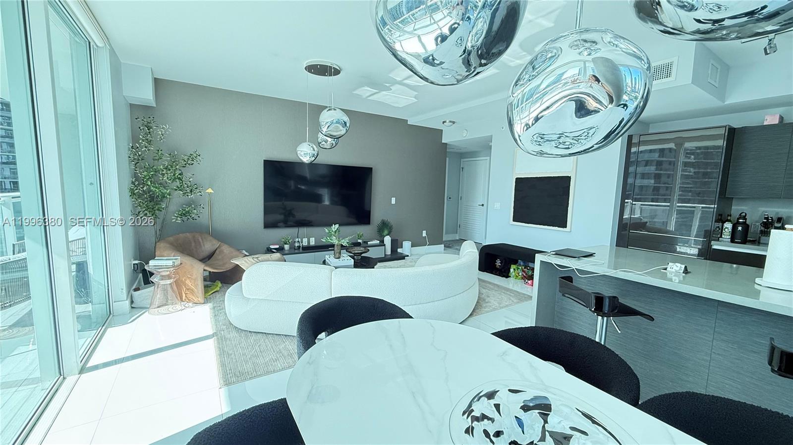 A modern living room features a curved white sofa, a large television, and floor-to-ceiling windows — 1080 Brickell Ave # 1801, Miami FL 33131, MIAMI, FL