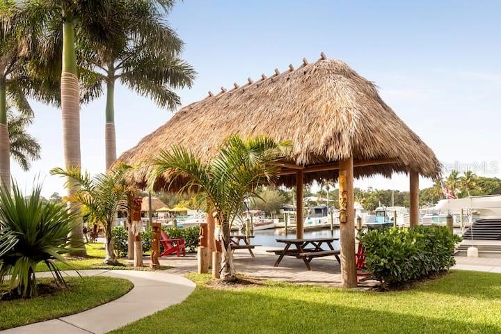 A thatched-roof tiki hut with picnic tables sits on a lawn near a marina — 7150 N TAMIAMI TRAIL UNIT S-613, SARASOTA, FL