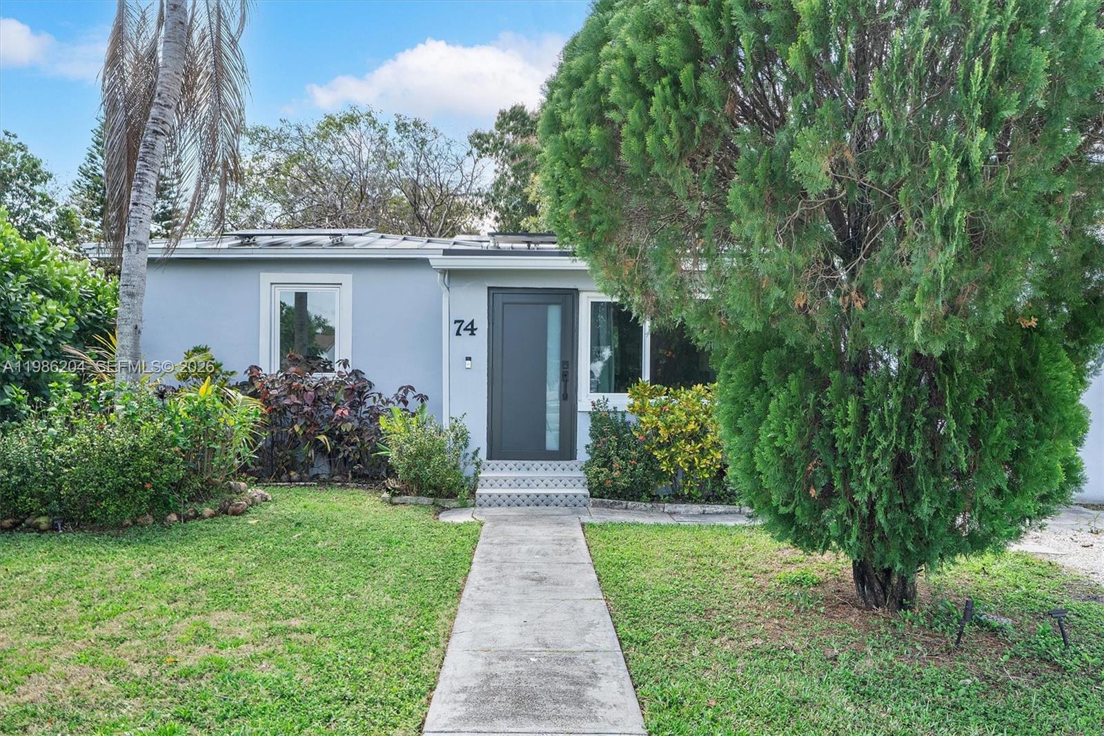 The exterior of a single-story house features a gray facade, a dark front door, and landscaping — 74 NW 117th St, Miami FL 33168, MIAMI, FL