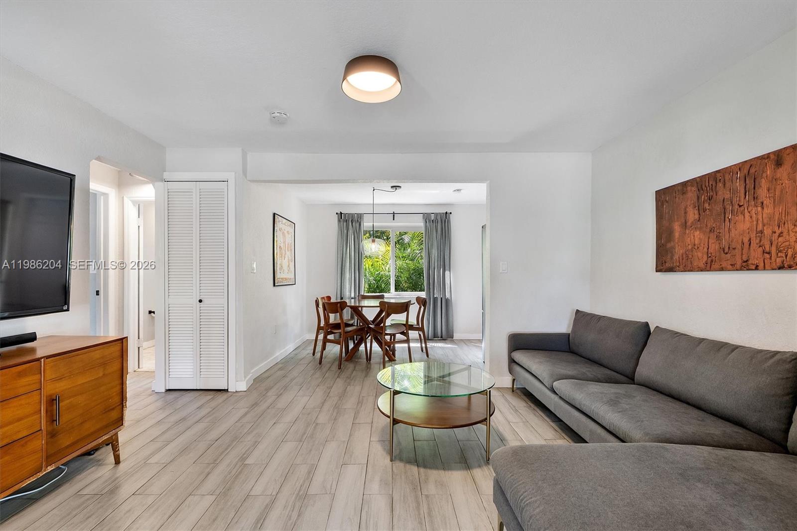 A living room with light wood-look flooring leads into a dining area with a window — 74 NW 117th St, Miami FL 33168, MIAMI, FL