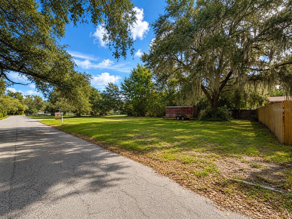 A grassy vacant lot sits beside a paved road with a trailer parked under trees — GASKINS STREET, LEESBURG, FL