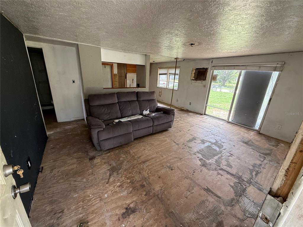 A living room features a grey sofa, unfinished plywood flooring, and a sliding glass door — 10360 SE 131ST LANE, OCKLAWAHA, FL