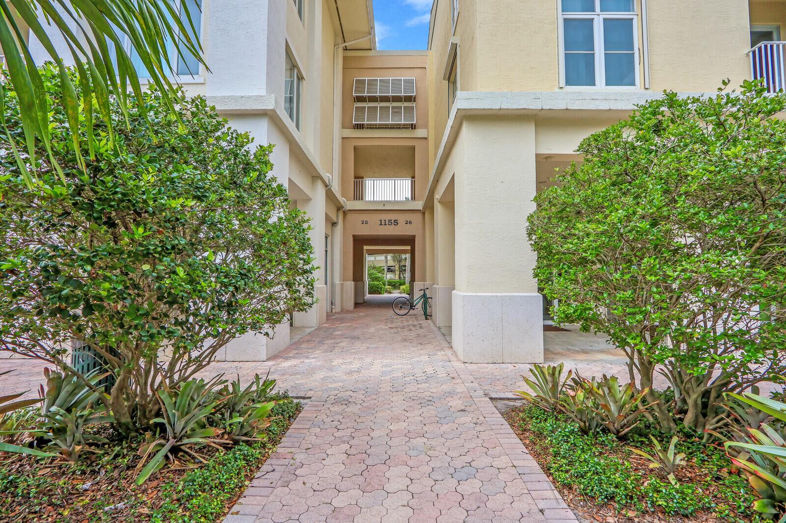 A paved walkway leads between two beige apartment buildings with lush green landscaping — 1155 Main Street # 216, Jupiter FL 33458, JUPITER, FL