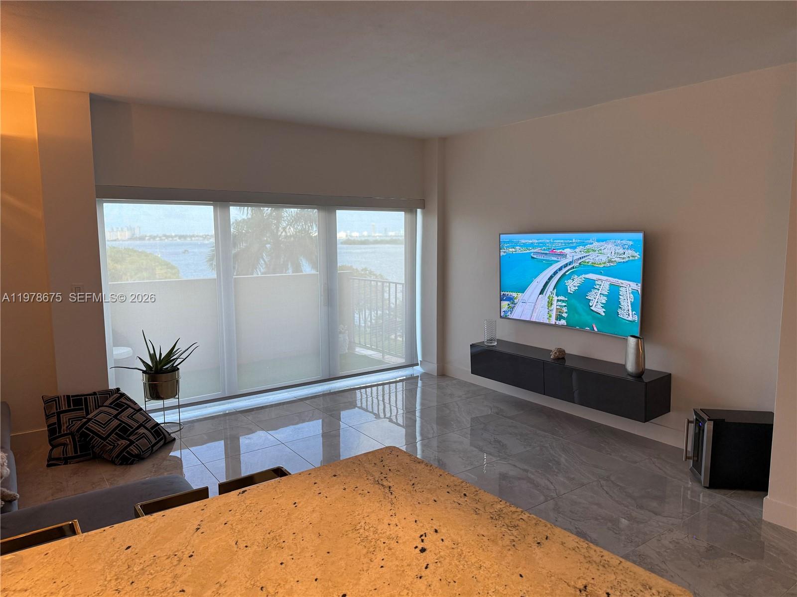 A living room features a floating console, wall-mounted television, tiled floors, and a balcony view — 880 NE 69th St # 5Q, Miami FL 33138, MIAMI, FL