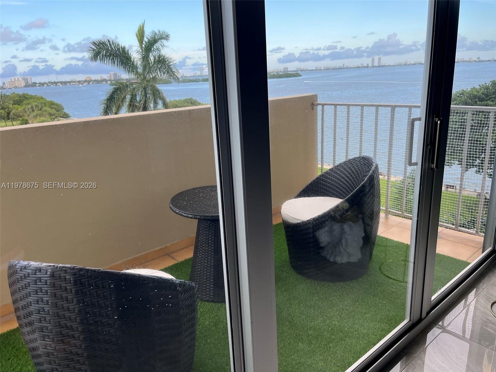 A balcony with artificial turf, two wicker chairs, and a small table overlooks the water — 880 NE 69th St # 5Q, Miami FL 33138, MIAMI, FL