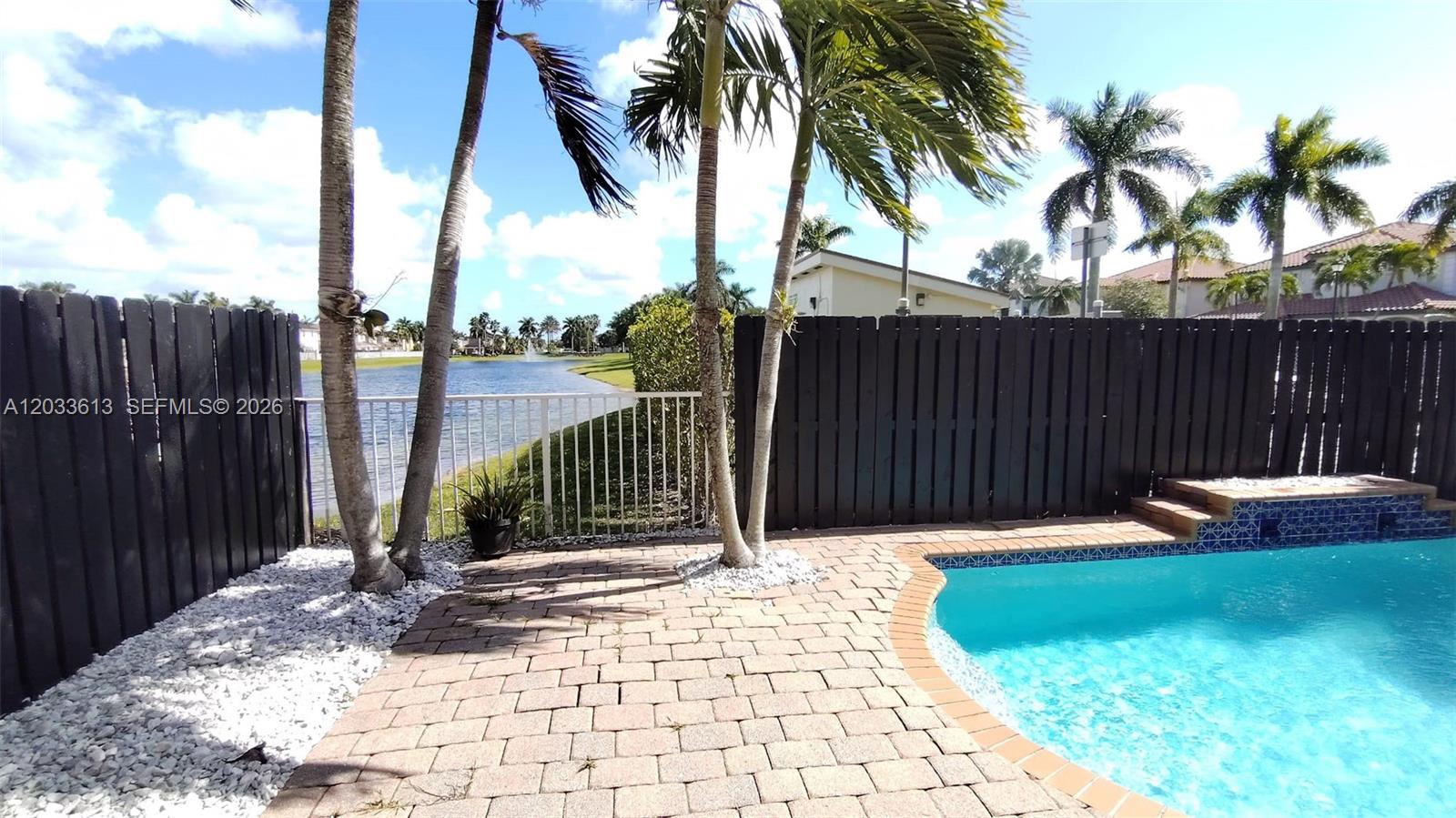 A swimming pool with a brick patio sits beside a lake and palm trees — 6743 SW 163rd Pl, Miami FL 33193, MIAMI, FL