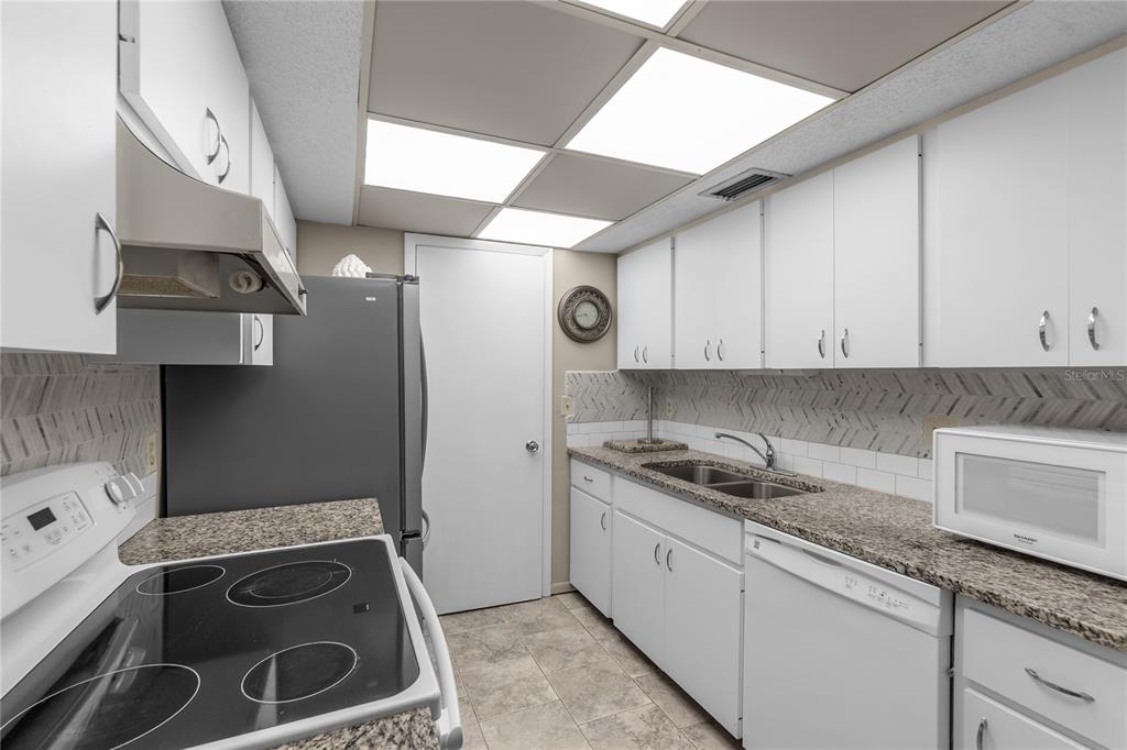 A galley kitchen features white cabinets, granite countertops, a herringbone backsplash, and stainless steel appliances — 4119 61ST AVENUE TERRACE W UNIT 102, BRADENTON, FL