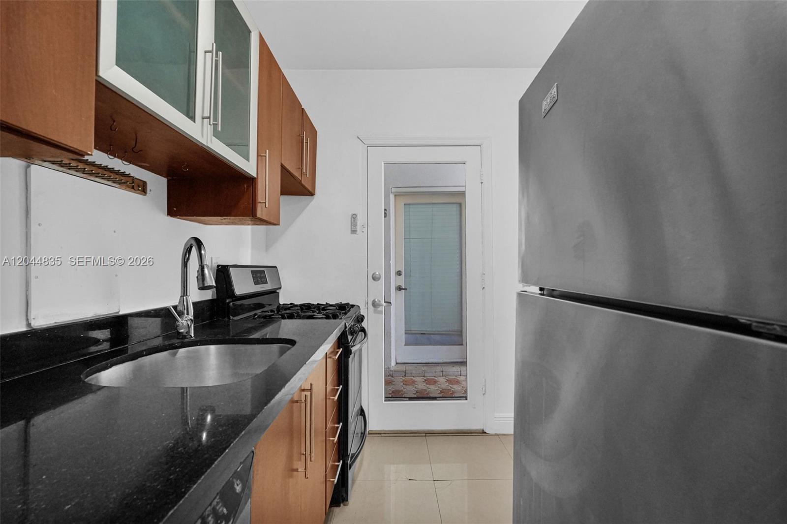 A galley kitchen features wood cabinets, black countertops, stainless steel appliances, and a glass-paneled door — 320 85th St # 4, Miami Beach FL 33141, MIAMI BEACH, FL