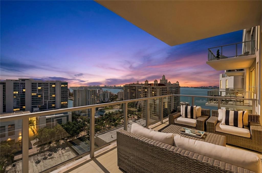 A balcony with wicker furniture overlooks a city skyline and water at sunset — 301 QUAY COMMONS UNIT 1807, SARASOTA, FL