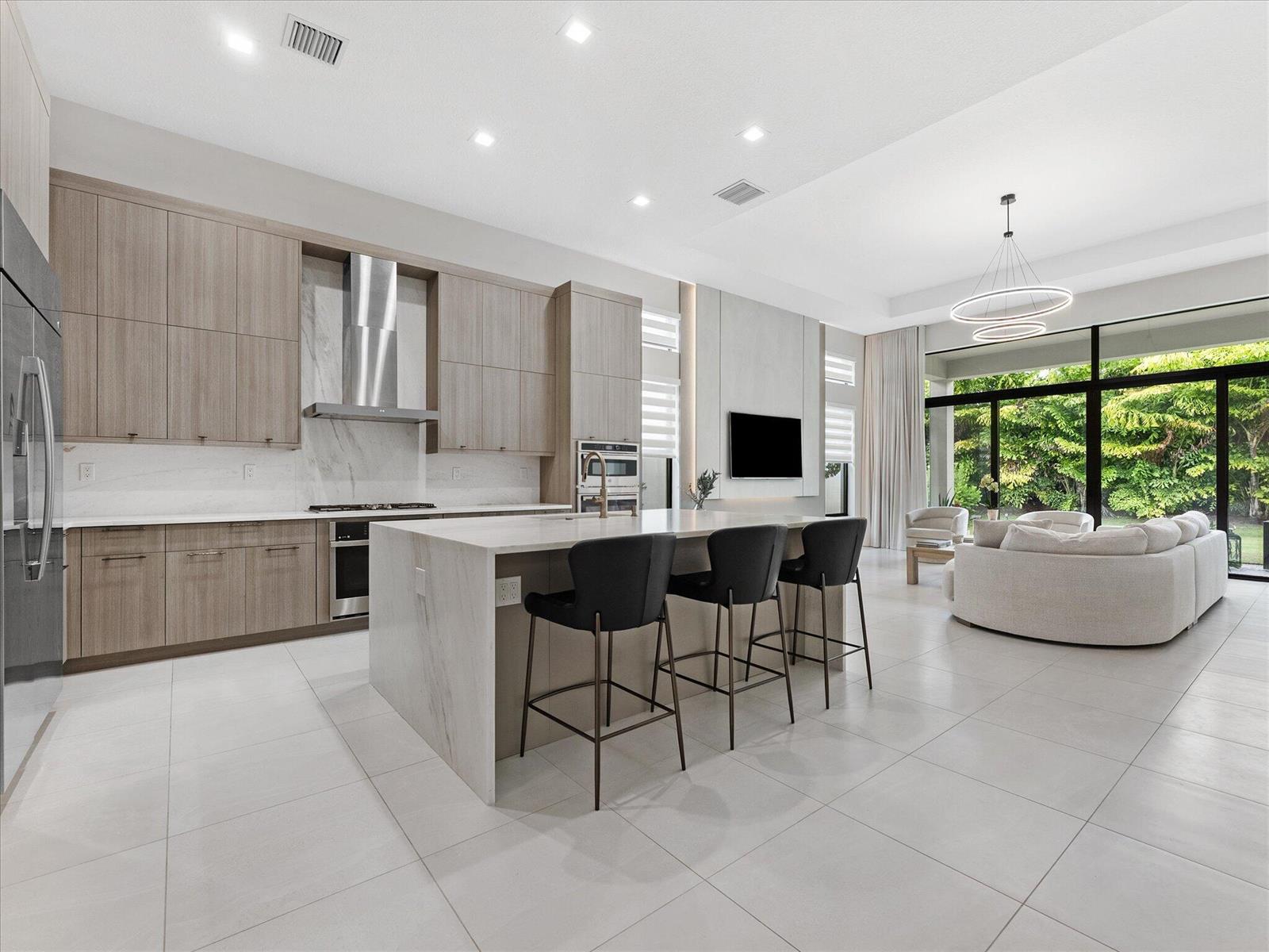A modern kitchen with light wood cabinets, a marble island, and an adjacent living area — 9273 Sunflower Meadow Road, Boca Raton FL 33434, BOCA RATON, FL