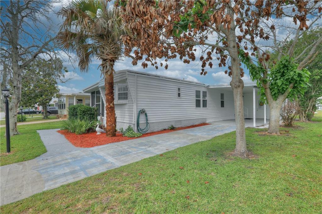 A white mobile home with a carport, a palm tree, and a red-mulched garden bed — BAREFOOT BAY, FL