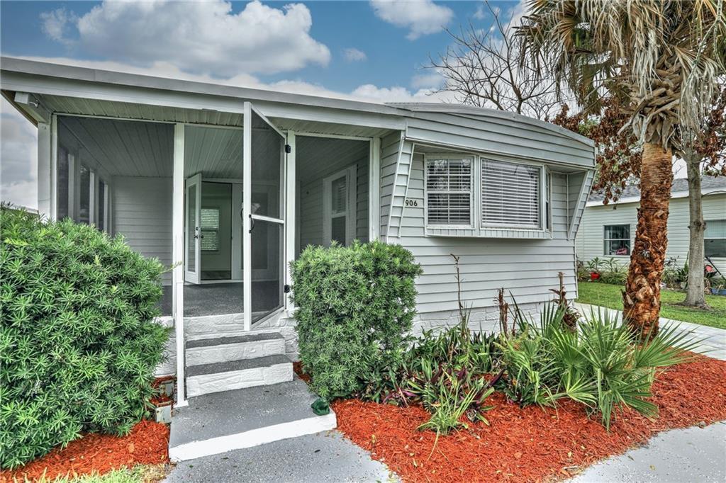 A light gray mobile home features a screened-in porch, white steps, and landscaped front yard — BAREFOOT BAY, FL