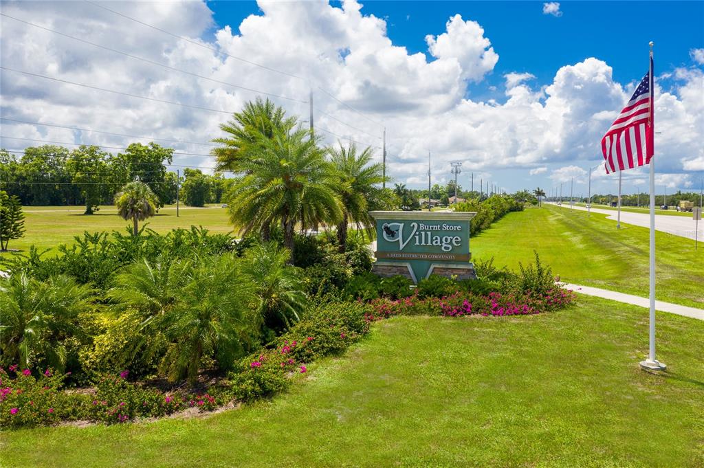A sign for Burnt Store Village stands in a landscaped area next to an American flag — 16216 QUESA DRIVE, PUNTA GORDA, FL