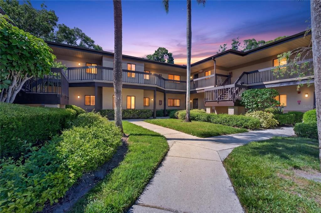 A two-story apartment complex exterior with a concrete walkway, green landscaping, and palm trees — 2691 SABAL SPRINGS CIRCLE UNIT 201, CLEARWATER, FL
