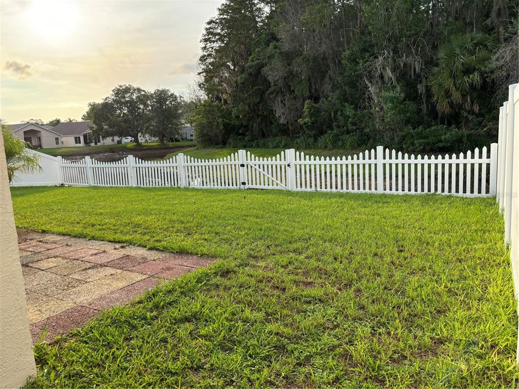 A grassy backyard features a white picket fence bordering a pond and wooded area — 4304 STONES RIVER COURT, NEW PORT RICHEY, FL
