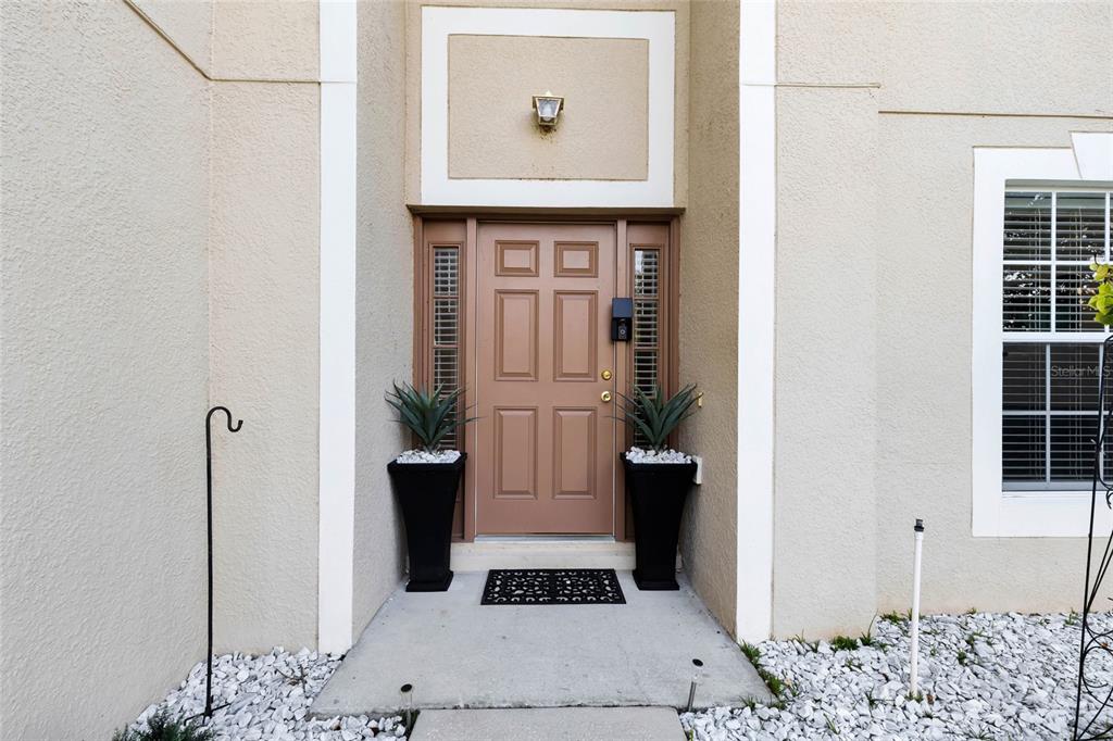 A tan front door with sidelights is flanked by two black planters on a porch — 1702 FOREST GLEN DRIVE, FRUITLAND PARK, FL