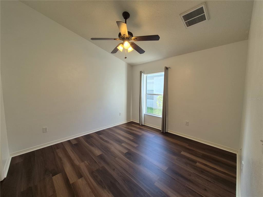 An empty room features dark wood-look flooring, white walls, a ceiling fan, and a window — 6833 RUNNER OAK DRIVE, ZEPHYRHILLS, FL